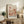 May include: Framed art print displayed on rustic wooden sideboard in warm-toned living room setting. Print features abstract botanical illustration with coral, pink and cream colors showing stylized mushrooms, flowers and organic shapes with polka dot patterns. Japanese text visible at bottom. Natural wood frame complements earth-toned decor including potted fern, ceramic vases, books, and beige upholstered furniture with throw pillows.
