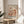 May include: Framed art print in natural wood frame sitting on round wooden side table next to beige upholstered chair. The poster features abstract botanical illustration with coral and pink mushrooms, flowers, and organic shapes in watercolor style, with "YAYOI KUSAMA" and "KYOTO" text at bottom. Soft natural lighting creates shadows on white wall, with plant leaves visible in corner.