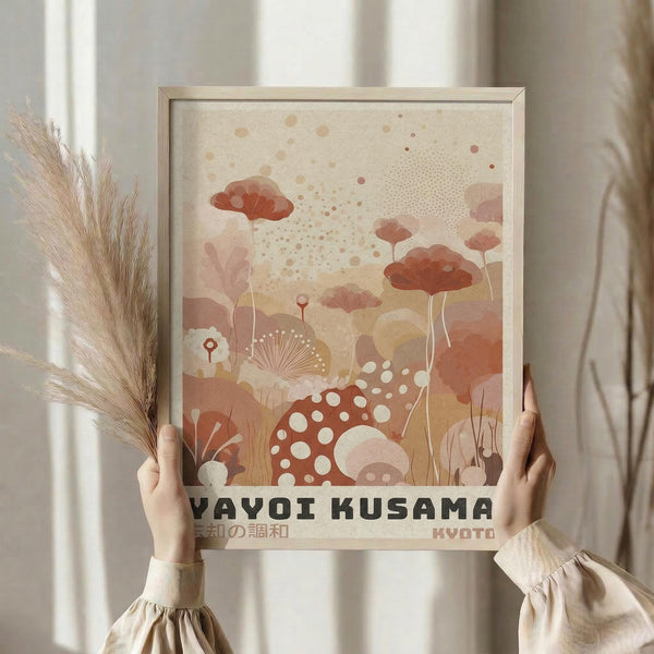 May include: Person holding framed abstract art print featuring warm terracotta and cream colored organic shapes, dots, and botanical elements including mushrooms and fan-like forms. Light wood frame against neutral curtained background with dried pampas grass visible on left side. Soft natural lighting creates calm, minimalist styling.