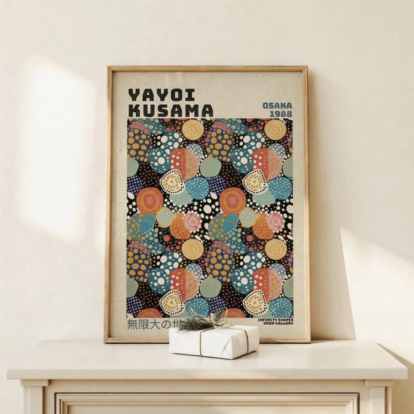 May include: Framed art print with colorful polka dot pattern in blues, oranges, and earth tones displayed on light wood shelf against cream wall. Print features overlapping circles with dotted textures in Kusama's signature style, showing "OSAKA 1988" text. Small wrapped gift box with twine sits beside the thin wooden frame.