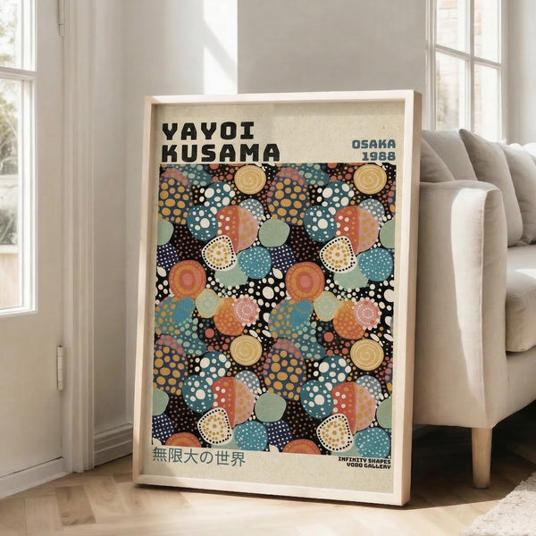 May include: Framed art print leaning against white wall next to beige upholstered chair in bright living room. Poster features colorful polka dot pattern with overlapping circles in blues, oranges, greens, and earth tones on black background. Text reads "YAYOI KUSAMA" and "OSAKA 1988" at top, Japanese characters at bottom. Light wood frame, natural lighting from large windows.