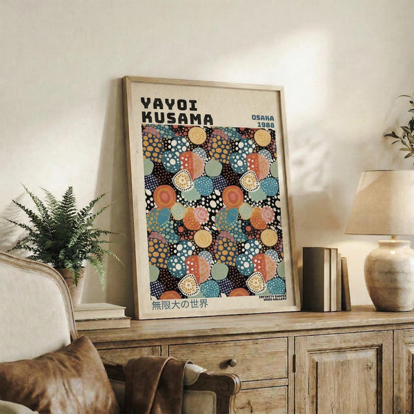 May include: Framed art poster with colorful polka dot pattern in blues, oranges, and earth tones displayed on rustic wooden sideboard. Room features neutral walls, potted fern, ceramic vase, stacked books, and brown leather armchair. Natural lighting creates warm, cozy living space atmosphere.