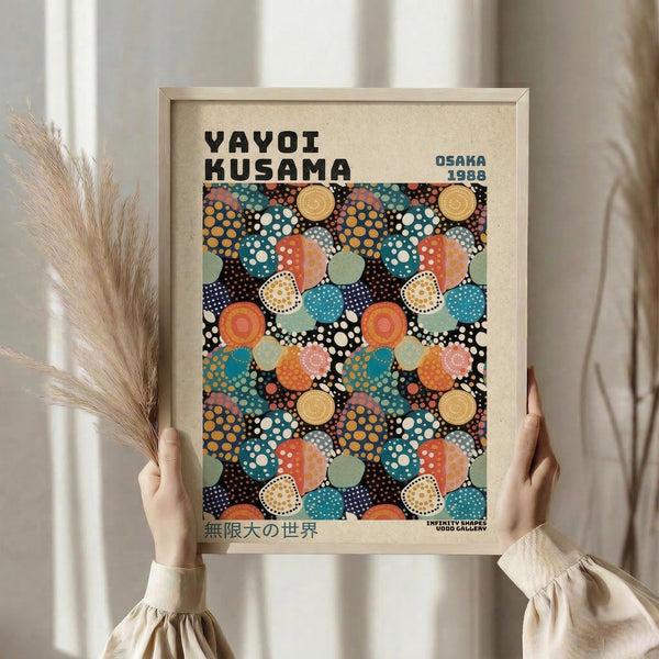 May include: Person holding framed poster featuring colorful polka dot pattern in blues, oranges, reds, and yellows on black background. Text reads "YAYOI KUSAMA" and "OSAKA 1988" with Japanese characters below. Light wood frame against neutral curtained background with dried pampas grass visible on left side.