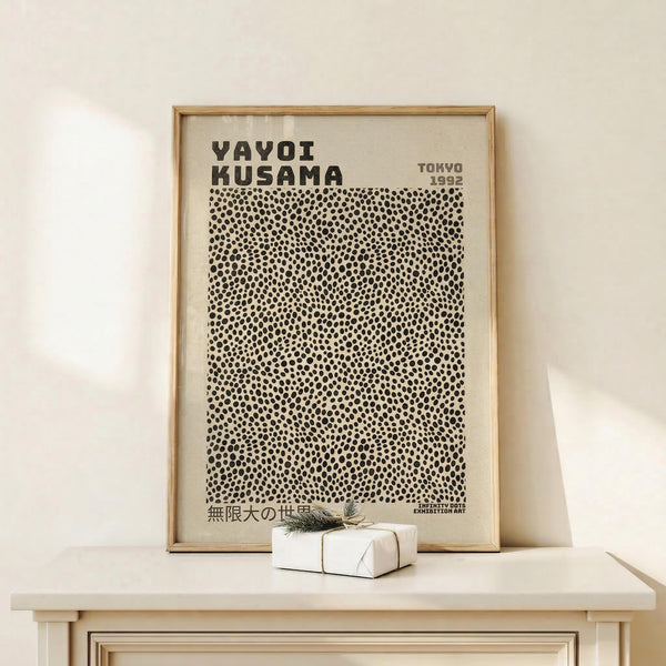 May include: Framed poster with black polka dot pattern on cream background, featuring "YAYOI KUSAMA" and "TOKYO 1992" text at top, displayed on light wood console table against white wall. Small wrapped gift box with twine and greenery sits beside the wooden frame.