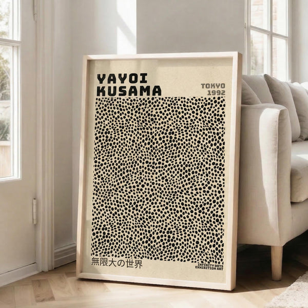 May include: Framed beige poster with black polka dot pattern leaning against white wall next to beige upholstered chair. Print features bold black text at top reading "YAYOI KUSAMA" and "TOKYO 1992" with dense field of irregular black dots covering most of the design. Japanese text appears at bottom. Natural wood frame, bright room with large windows casting soft shadows on wooden floor.