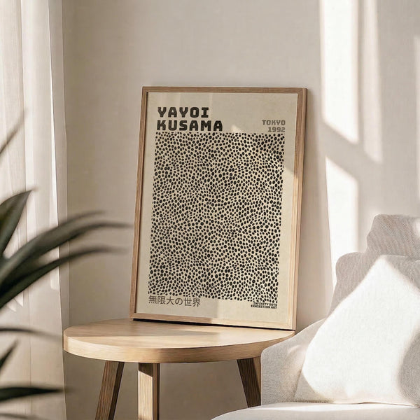 May include: Framed poster with cream background featuring black polka dot pattern and bold text reading "YAYOI KUSAMA" and "TOKYO" at top, Japanese characters at bottom. Natural wood frame sits on round wooden side table next to beige upholstered chair. Soft natural lighting creates window shadows on white wall, with green plant leaves visible in corner.