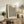 May include: Framed cream-colored poster with black polka dot pattern and bold text reading "YAYOI KUSAMA" and "TOKYO 1992" displayed on rustic wooden sideboard. Room setting features neutral tones, potted plants including fern and olive branches, stacked books, ceramic vase, and brown leather armchair. Natural lighting creates warm, cozy atmosphere with white walls.