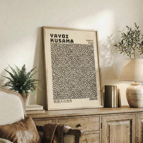 May include: Framed cream-colored poster with black polka dot pattern and bold text reading "YAYOI KUSAMA" and "TOKYO 1992" displayed on rustic wooden sideboard. Room setting features neutral tones, potted plants including fern and olive branches, stacked books, ceramic vase, and brown leather armchair. Natural lighting creates warm, cozy atmosphere with white walls.