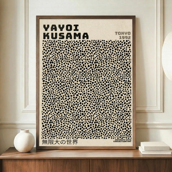 May include: Framed exhibition poster with bold black text "YAYOI KUSAMA" and dense pattern of black polka dots on cream background, displayed on wooden sideboard next to white ceramic vase and books. Warm neutral room with paneled walls and natural lighting creates minimalist modern aesthetic.
