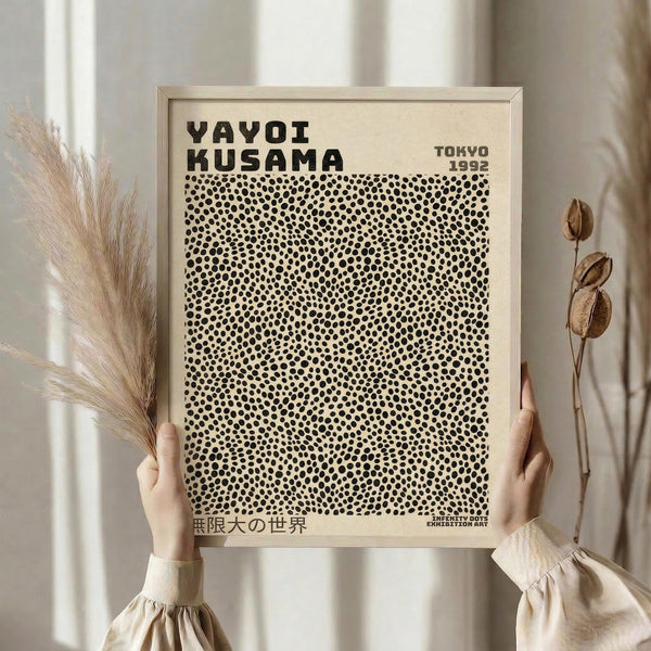 May include: Hands holding a framed art print featuring black polka dots on cream background with "YAYOI KUSAMA" and "TOKYO 1992" text, displayed in minimalist setting with dried pampas grass and seed pods, soft natural lighting, neutral beige tones throughout scene.