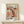 May include: Framed poster with abstract organic shapes in coral pink, cream, and black with polka dot patterns, displayed on white wooden shelf against cream wall. Text reads "YAYOI KUSAMA" and "TOKYO 1998" with Japanese characters below. Small wrapped gift box with twine sits beside the natural wood frame.