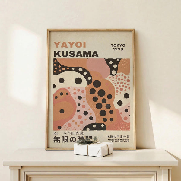 May include: Framed poster with abstract organic shapes in coral pink, cream, and black with polka dot patterns, displayed on white wooden shelf against cream wall. Text reads "YAYOI KUSAMA" and "TOKYO 1998" with Japanese characters below. Small wrapped gift box with twine sits beside the natural wood frame.