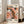 May include: A framed poster with abstract art in warm coral, pink, and orange tones featuring black and white polka dot patterns. The artwork displays text reading "YAYOI KUSAMA" and "TOKYO 1998" with Japanese characters below. The white wooden frame leans against a bright white wall next to large windows casting natural light. A beige upholstered sofa sits nearby on light wooden flooring with a white area rug.