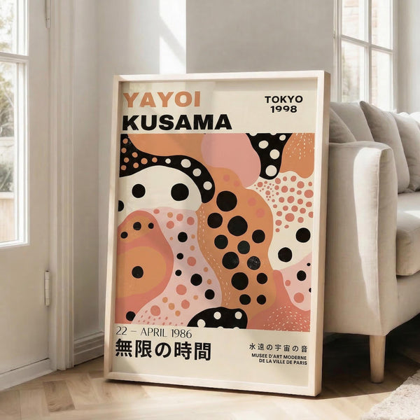 May include: A framed poster with abstract art in warm coral, pink, and orange tones featuring black and white polka dot patterns. The artwork displays text reading "YAYOI KUSAMA" and "TOKYO 1998" with Japanese characters below. The white wooden frame leans against a bright white wall next to large windows casting natural light. A beige upholstered sofa sits nearby on light wooden flooring with a white area rug.