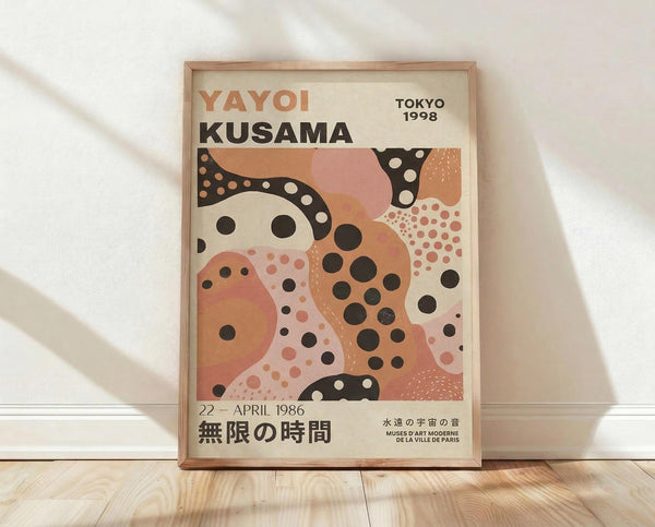 May include: Framed exhibition poster featuring abstract organic shapes in coral, peach, and cream tones with black and white polka dot patterns. Text reads "Yayoi Kusama Tokyo 1998" and "22 - April 1986" with Japanese characters. Natural wood frame leaning against white textured wall on light wooden floor with soft shadows.