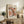 May include: Framed poster with abstract organic shapes in coral, peach, and cream tones featuring black polka dots, displayed on rustic wooden console alongside potted fern, ceramic vases, and books in neutral-toned living room setting