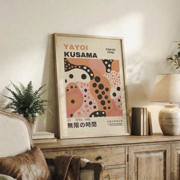 May include: Framed poster with abstract organic shapes in coral, peach, and cream tones featuring black polka dots, displayed on rustic wooden console alongside potted fern, ceramic vases, and books in neutral-toned living room setting