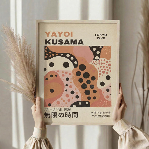 May include: Person holding framed abstract art poster with polka dot patterns in coral, peach, and black colors. Features Japanese text and exhibition details. Light wood frame, neutral background with decorative dried pampas grass visible on left side.