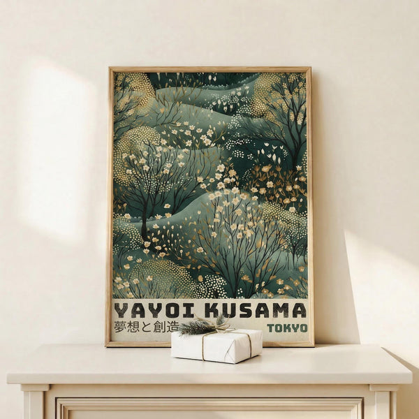 May include: Framed poster with green and cream botanical illustration featuring layered plants, flowers, and dotted patterns in an organic, nature-inspired design. Text reads "YAYOI KUSAMA" and "TOKYO" in bold black letters with Japanese characters. Light wood frame sits on white wooden surface next to small wrapped gift box with twine and dried sprig against cream wall.