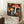 May include: Framed vintage-style exhibition poster featuring colorful mushrooms and fungi on black starry background, leaning against white wall corner next to small wooden side table with white coffee cup and saucer, cozy textured throw blanket visible at bottom right, natural window lighting creating soft shadows on white walls and fluffy area rug below.