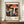 May include: Framed vintage exhibition poster featuring colorful mushrooms and fungi on black dotted background, displayed on wooden sideboard next to white ceramic vase and books. Poster shows red spotted toadstools, orange and yellow mushrooms with white stems, blue foliage elements. Text reads "28. APRIL - 14. MAI 1984" and "YAYOI KUSAMA" with "MUSÉE D'ART MODERNE DE LA VILLE DE PARIS" below. Warm beige wall background with white trim molding visible.
