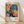 May include: Framed exhibition poster featuring vibrant floral artwork with polka dot patterns in pink, blue, yellow, and black. Shows stylized flowers and organic shapes with dotted textures characteristic of pop art style. Text reads "YAYOI KUSAMA" with Japanese characters and museum details, dated 1984. Natural wood frame leaning against white wall on light wooden floor with soft shadows.