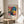 May include: Framed colorful floral art poster displayed on light wooden round stool next to beige upholstered chair. Artwork features vibrant flowers in pink, blue, yellow with polka dot patterns against black background. Exhibition poster shows museum text and Japanese characters. Natural sunlight streams across white wall creating soft shadows from nearby plant leaves.