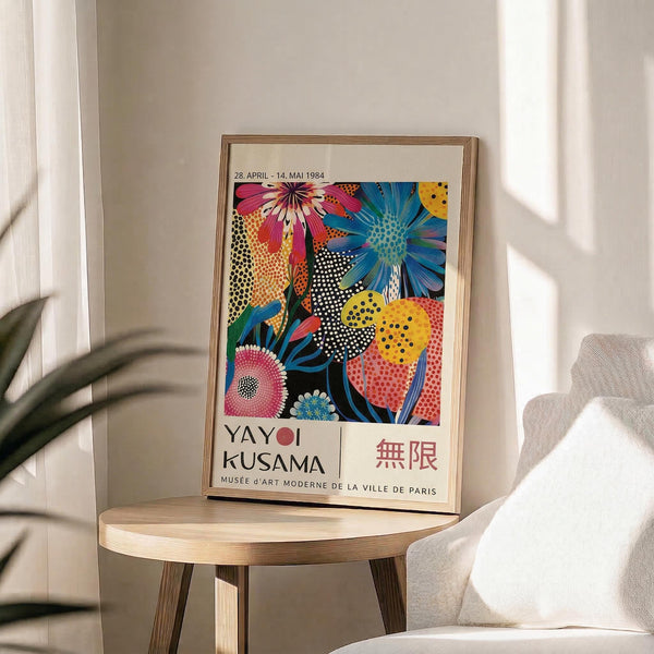 May include: Framed colorful floral art poster displayed on light wooden round stool next to beige upholstered chair. Artwork features vibrant flowers in pink, blue, yellow with polka dot patterns against black background. Exhibition poster shows museum text and Japanese characters. Natural sunlight streams across white wall creating soft shadows from nearby plant leaves.