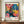 May include: Framed vintage exhibition poster featuring vibrant pop art flowers in pink, blue, yellow and orange with polka dot patterns, displayed on wooden sideboard next to white ceramic vase and books against cream wainscoted wall. Poster shows bold floral design with Japanese and French text dated 1984.