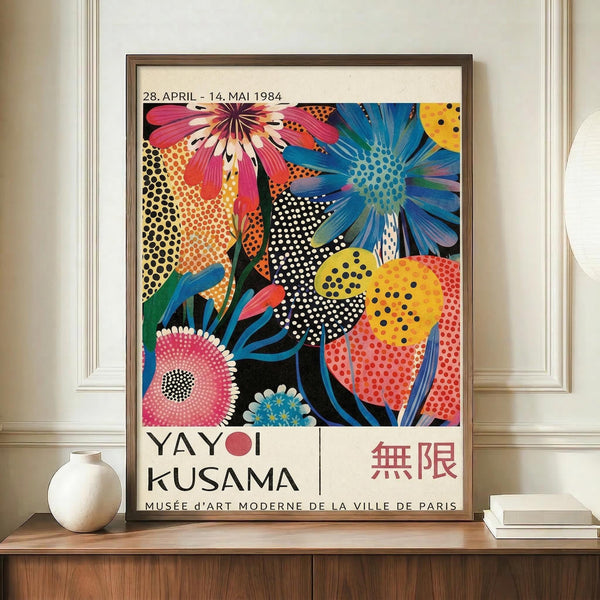 May include: Framed vintage exhibition poster featuring vibrant pop art flowers in pink, blue, yellow and orange with polka dot patterns, displayed on wooden sideboard next to white ceramic vase and books against cream wainscoted wall. Poster shows bold floral design with Japanese and French text dated 1984.