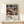 May include: Framed colorful abstract art poster with swirling patterns in blues, oranges, purples and yellows, featuring organic flowing shapes and polka-dotted elements. Text reads "YAYOI KUSAMA" with Japanese characters and French museum text below. Natural wood frame sits on light wooden surface against cream wall, with small wrapped gift box beside it.