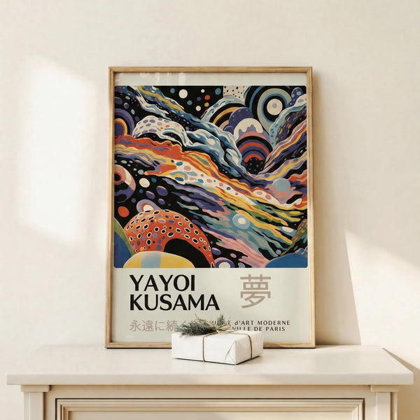 May include: Framed colorful abstract art poster with swirling patterns in blues, oranges, purples and yellows, featuring organic flowing shapes and polka-dotted elements. Text reads "YAYOI KUSAMA" with Japanese characters and French museum text below. Natural wood frame sits on light wooden surface against cream wall, with small wrapped gift box beside it.