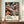 May include: Framed poster with vibrant abstract swirling patterns in orange, purple, blue and white, featuring polka-dotted organic shapes against flowing curved lines. Text reads "YAYOI KUSAMA" with Japanese characters and "MUSÉE D'ART MODERNE DE LA VILLE DE PARIS" below colorful artwork. Poster displayed on wooden sideboard next to white ceramic vase and stacked books in elegant room with cream walls and white trim molding.