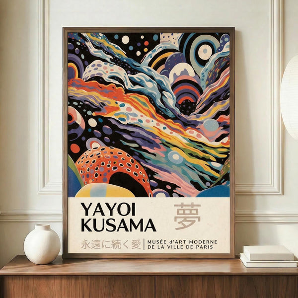 May include: Framed poster with vibrant abstract swirling patterns in orange, purple, blue and white, featuring polka-dotted organic shapes against flowing curved lines. Text reads "YAYOI KUSAMA" with Japanese characters and "MUSÉE D'ART MODERNE DE LA VILLE DE PARIS" below colorful artwork. Poster displayed on wooden sideboard next to white ceramic vase and stacked books in elegant room with cream walls and white trim molding.