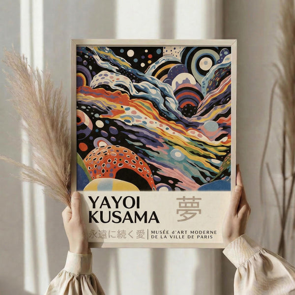May include: Person holding framed art print featuring colorful abstract swirling patterns in orange, blue, purple, and cream with polka dots and flowing organic shapes. Text reads "YAYOI KUSAMA" with Japanese characters and "MUSÉE d'ART MODERNE DE LA VILLE DE PARIS". Light wooden frame, neutral beige setting with dried pampas grass visible to the left.
