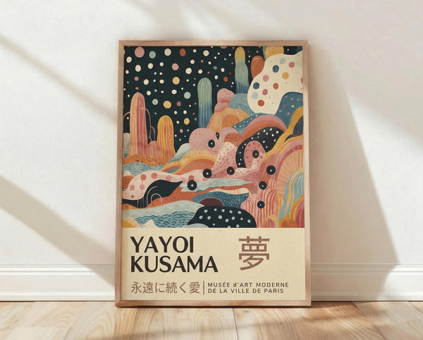 May include: Framed art print leaning against white wall on light wood floor. Colorful abstract artwork features flowing organic shapes, cacti, rainbow arcs, and polka dot patterns in coral, teal, yellow, and navy. Dark starry sky transitions to whimsical landscape with dotted textures. Text reads "YAYOI KUSAMA" with Japanese characters and museum attribution below. Natural wood frame, soft lighting with shadows.