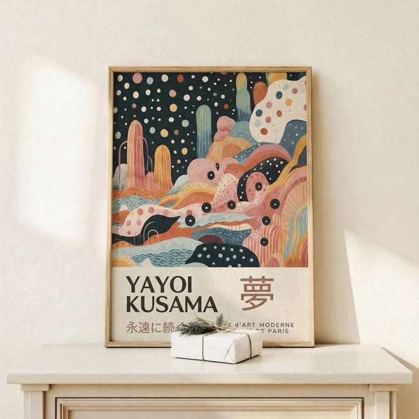 May include: Framed art print featuring abstract desert landscape with colorful cacti, rolling hills, and starry night sky in coral, sage green, and navy tones. Shows "YAYOI KUSAMA" text with Japanese characters below. Wooden frame displays on white floating shelf next to small wrapped gift with evergreen sprig against cream wall.