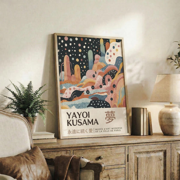 May include: Framed poster with abstract landscape featuring colorful cacti, flowing organic shapes, and polka dot patterns against a starry night sky. Japanese text visible at bottom. Displayed on rustic wooden sideboard beside fern plant, ceramic vase, and books in modern living room with beige armchair and neutral walls.