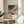May include: Framed art poster displaying abstract colorful illustration with polka dot sky, desert cacti, and flowing organic shapes in pink, orange, blue, and green tones. Features "YAYOI KUSAMA" text and Japanese characters below artwork. Wooden frame sits on round natural wood side table next to beige upholstered chair. Soft natural lighting creates shadows on white wall. Green plant leaves visible in corner.