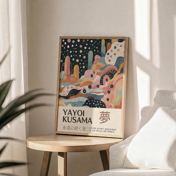 May include: Framed art poster displaying abstract colorful illustration with polka dot sky, desert cacti, and flowing organic shapes in pink, orange, blue, and green tones. Features "YAYOI KUSAMA" text and Japanese characters below artwork. Wooden frame sits on round natural wood side table next to beige upholstered chair. Soft natural lighting creates shadows on white wall. Green plant leaves visible in corner.