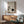 May include: Framed artwork displaying a minimalist bonsai tree with drooping branches and a large dark circular moon against a muted beige textured background, shown in a modern living room setting with a sage green mid-century armchair, wooden sideboard, white ceramic table lamp, and natural sunlight casting shadows on white walls.