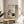 May include: Framed botanical art print displayed on round wooden side table next to beige upholstered chair. Artwork features minimalist zen design with bonsai tree branch, dark circular moon shape, and hanging ornament against neutral background. Natural wood frame complements warm sunlit room with plant shadows on cream walls.