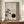May include: A framed minimalist artwork featuring a stylized bonsai tree with delicate green foliage extending from the left side, a large black circle resembling a moon, and a small dark spherical ornament hanging below. The piece has a neutral beige textured background and is displayed on a wooden sideboard alongside a cream ceramic vase and white books in an elegant room setting.