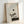 May include: Framed minimalist artwork leaning against white wall, featuring zen-style composition with delicate pine tree branch, geometric shapes including black and gray circles connected by thin line, and abstract rectangular forms in muted earth tones, all set within light wooden frame on neutral surface