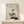 May include: Framed minimalist art print featuring a delicate bonsai tree with geometric shapes - a gray textured circle, black solid circle, and white square - arranged above the tree against a neutral background. The print is displayed in a light wood frame on a pale wooden surface alongside a white gift box tied with twine and evergreen sprig. Soft natural lighting creates gentle shadows on the cream-colored wall behind.