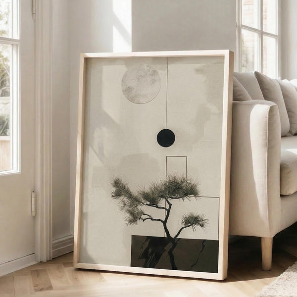 May include: Framed minimalist artwork featuring a delicate bonsai pine tree with curved trunk and needle clusters against a soft beige background, accompanied by geometric shapes including a gray circle, black circle, and outlined rectangle. The piece is displayed in a light wood frame, leaning against a white wall next to a beige upholstered sofa in a bright, sunlit room with large windows and wooden floors.