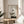 May include: Framed zen-style artwork featuring minimalist bonsai tree with geometric shapes including circles and rectangles in neutral tones, displayed on round wooden side table next to beige upholstered chair. Soft natural lighting creates shadows on cream wall, with plant leaves visible in corner of serene modern interior setting.