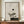 May include: Framed zen-inspired artwork featuring a minimalist bonsai tree with pine needles, geometric shapes including a gray circle and black dot against a textured beige background with rectangular elements. The wooden-framed print sits on a walnut credenza alongside a cream ceramic vase and white books in a neutral-toned room with panel molding.
