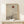 May include: Framed minimalist artwork leaning against white wall on light wood console table, featuring bare tree silhouette against layered mountain landscape with rust-colored sun on beige textured background, natural wood frame, small white wrapped gift with greenery beside it, soft natural lighting