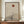 May include: Framed minimalist artwork featuring a bare black tree silhouette against beige textured background with terracotta sun and layered mountain shapes, displayed on walnut wood sideboard beside white ceramic vase and stacked books in neutral-toned room with white paneled walls.